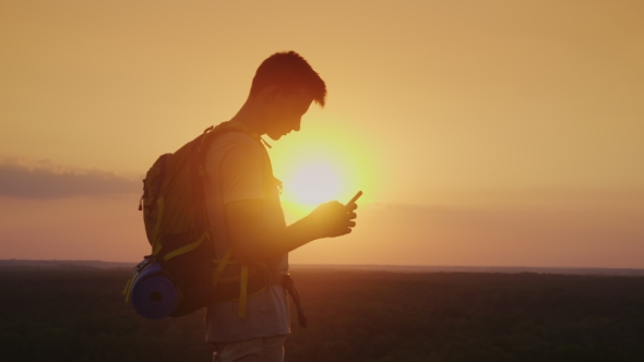 Always and Everywhere in Touch. A Traveler with a Backpack Uses a Smartphone. Silhouette on Sunset
