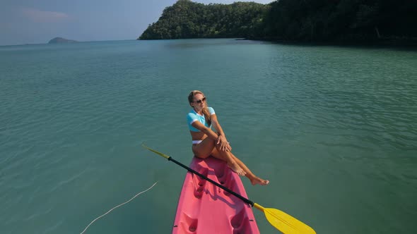 Pretty Sportive Blonde Lady in Stylish Swimsuit Sails on Pink Plastic Canoe Along Azure Ocean Bay at