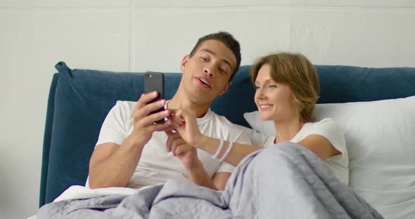 Mixed Race Couple Relaxing in Bed, Using Smartphone, Gadget