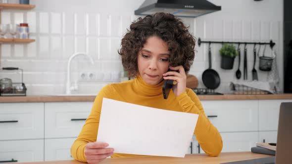 Businesswoman Talking on Mobile Phone with Financial Report in Hand in Kitchen
