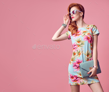 y Redhead Model in fashion pose. Trendy Floral Dress, Stylish wavy hairstyle, fashion Sunglasses, Luxury summer Clutch. Playful summer Girl on Pink