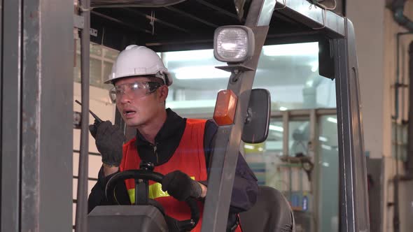 Asian male warehouse driver talking to walkie-talkie with colleague while driving forklift car