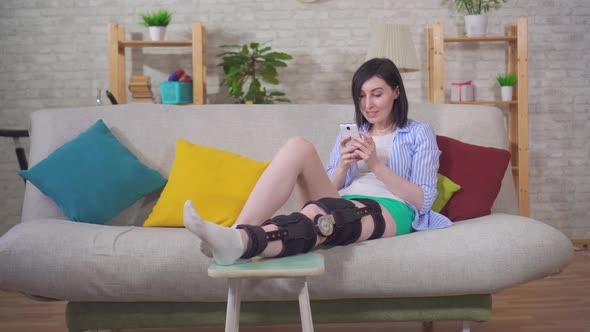 Female with the Phone After Injury Knee Bend Orthosis Knee Brace Lies on the Couch at Home