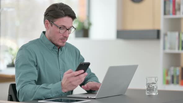 Middle Aged Man Using Smartphone While Working on Laptop