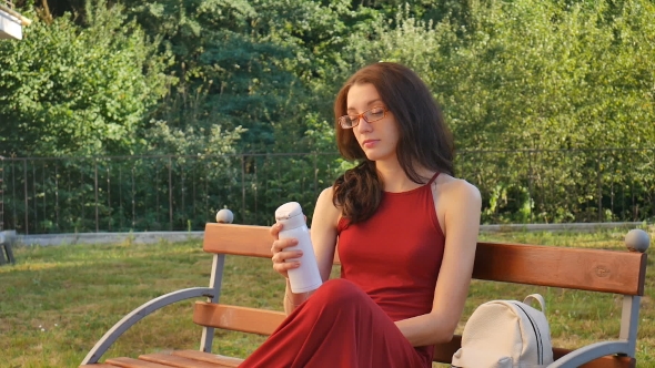 Cute Female Teenager in Dark Red Long Dress and Eyeglasses Drinking Something from Thermo Cup