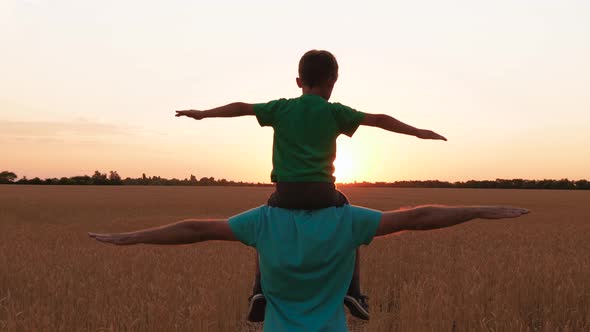 A Small Boy Sits on His Father's Neck, Imitating the Flight of an Airplane Over a Wheat Field