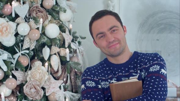 Smiling Young Man Holding a Book Standing Next To a Christmas Tree