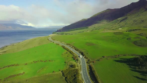 Aerial video of a Van driving on a Coastal road with green paddocks, blue ocean, and mountains.