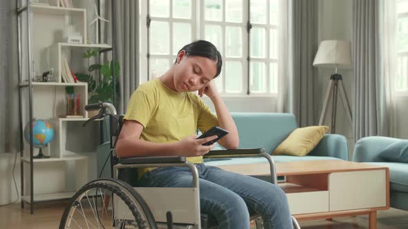 Bored Asian Kid Girl Sitting In A Wheelchair While Using Smart Phone At Home