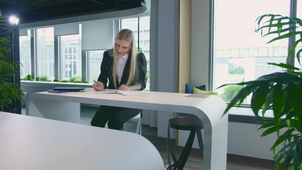 Elegant Woman Working in Stylish Office. Modern Blond Woman in Trendy Suit Sitting at Table in Light