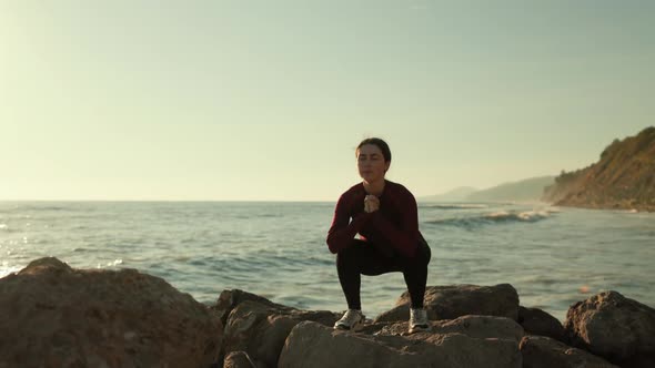 A sporty young woman squats on a wild beach. There is a stormy sea in the background.