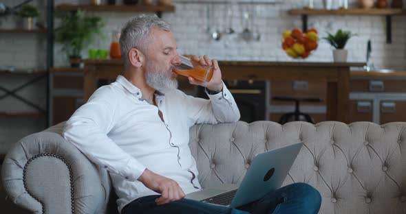 Handsome Mature Bearded Man with Grey Hair Working on Laptop Computer and Drinking Orange Juice