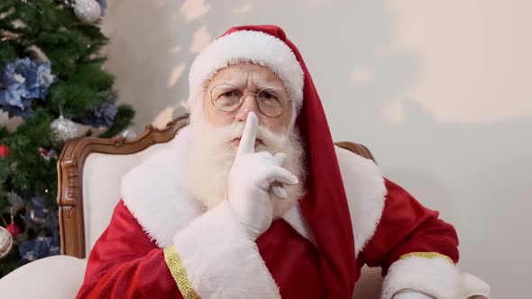 Santa Claus with red sack keeping forefinger by his mouth and looking at camera. Shh!