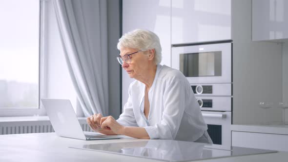 Depressed pensioner businesswoman types on grey laptop takes glasses off and wipes tears with hand