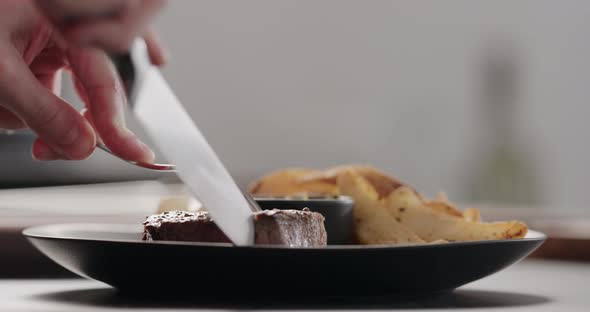 Slow Motion Man Slicing Beef Steak on Black Plate with Roasted Potatoes