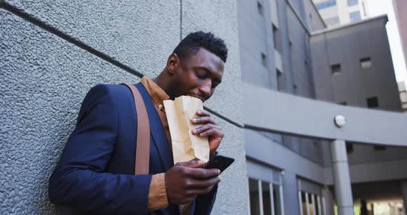 African american businessman eating sandwich using smartphone in city street