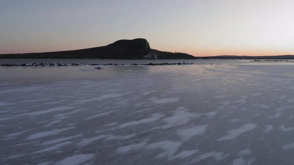 Sliding above snowy patterns in ice on the shore of a lake at dawn with mountain in distance AERIAL