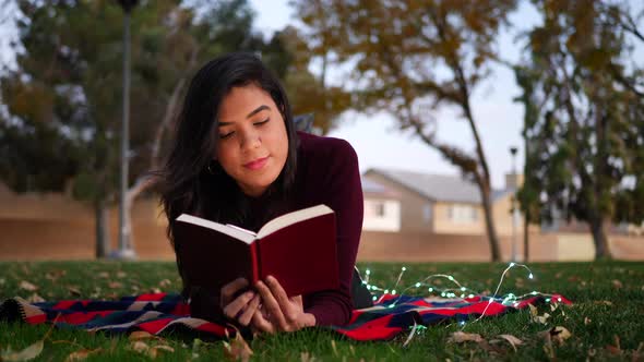 Close up of a young hispanic woman reading the pages of a story book laying down outdoors in the par