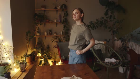 A Beautiful Smiling Young Girl Stands By a Wooden Table in a Room with a Blinking Garland and Many