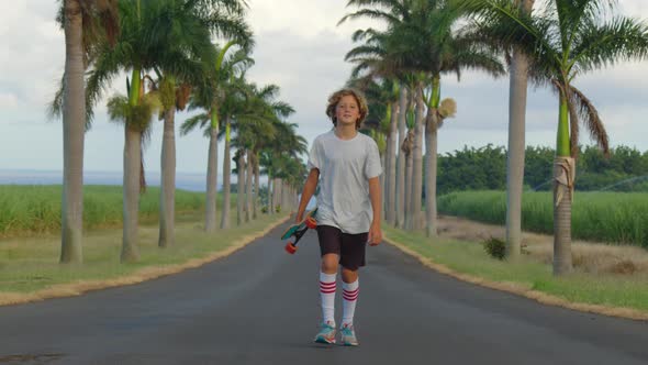 A Teenager with Long Hair Rides a Skateboard Along a Beautiful Road with Palm Trees