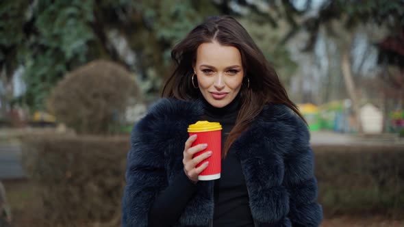 Portrait of glamorous girl with plastic coffee cup. Attractive young model in luxury fur coat standi