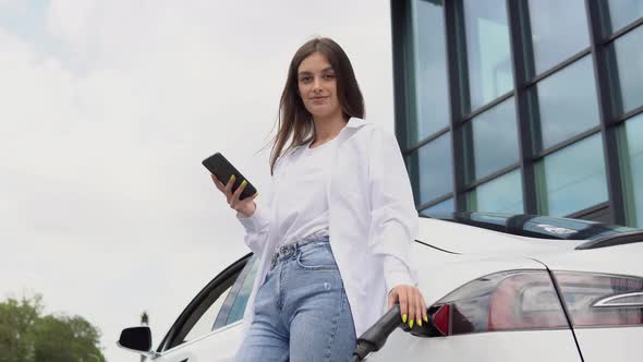 Young Woman is Standing Near the Electric Car and Looks at the Smart Phone