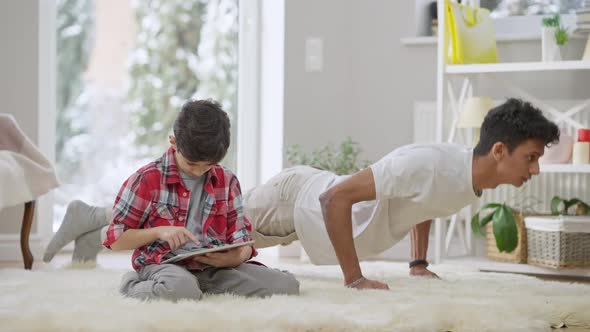 Absorbed Middle Eastern Little Boy Using Tablet As Teenage Brother Doing Pushups at Home