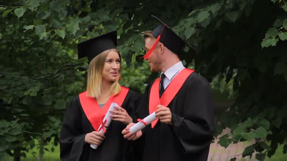 Smiling Couple of Graduates Walking in Park, Holding Diplomas and Talking