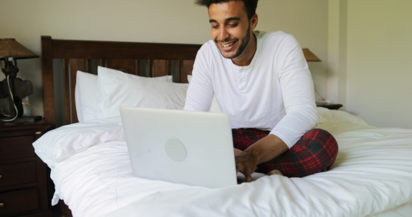 Pondering Man Using Laptop Computer Sitting On Bed Hispanic Guy Type Chatting Online In Bedroom