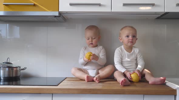 Two Twin Baby Girls Play on Kitchen Table Top with Lemons Patiently Waiting for Food to Be Cooked