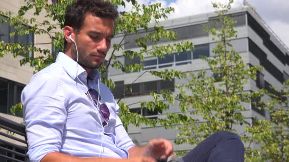 A Young Handsome Businessman Sits on a Bench and Listens To Music on a Smartphone, Office Building