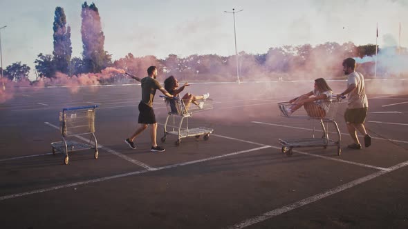 Young Fellows Holding Smoke Bombs Spinning and Racing on Shopping Carts with Girlfriends at Deserted