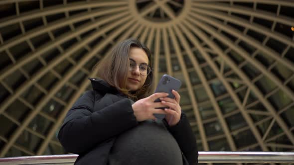 A Young Pregnant Woman in Glasses and a Jacket Stands with a Phone in Her Hands