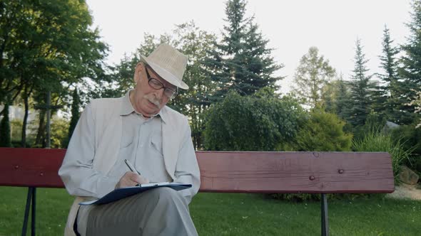 Elegant Senior Man in Hat and Suit Sitting on Park Bench and Writing on Notebook