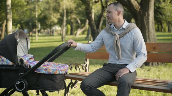 Portrait of Happy Young Father Rocking Baby Stroller and Smiling at Camera. Positive Caucasian