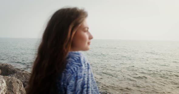 A Woman Smiles Looking Into the Distance Sitting on Boulders on the Seashore