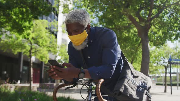 African american senior man wearing face mask using smartphone while leaning on his bicycle on the r