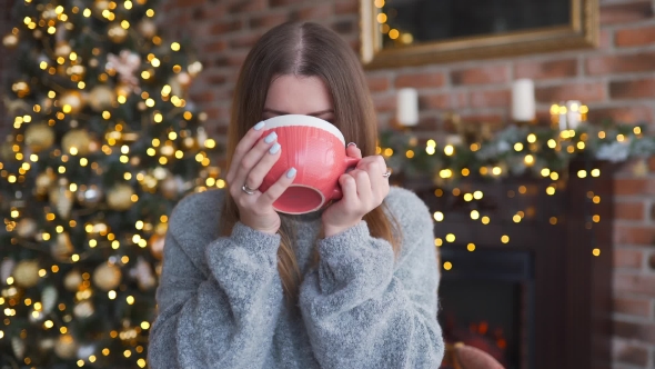 Happy Smiling Woman in the Sweater Drinks Hot Chocolate Near the Shining Christmas Tree at Her