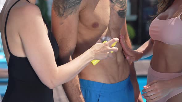 Hands of Two Unrecognizable Women Spraying Spreading Sunscreen on Muscular Man in Sunshine