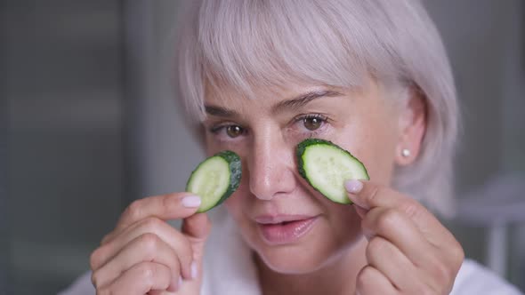 Closeup Face of Mature Woman with Grey Hair and Brown Eyes Putting Cucumber on Eye Skin Smiling