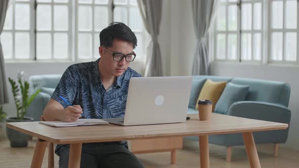 Asian Man Using Laptop Computer And Writing On Notebook At Home
