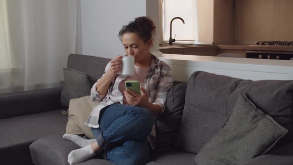 Woman Using Smartphone Drinking Hot Beverage with White Mug Indoors