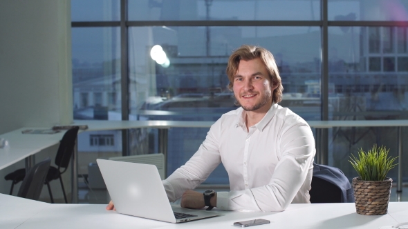 Portrait of Happy Young Businessman with Laptop Sitting By Desk in Office.