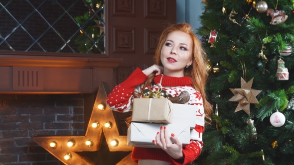 Cute Redhead Girl in the Red Sweater Near Christmas Tree with Gifts in Hand.