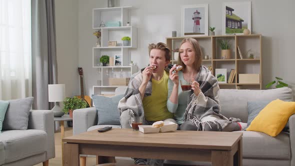 Young Couple Watching Tv and Drinking Coffee with Donuts Hiding in a Blanket