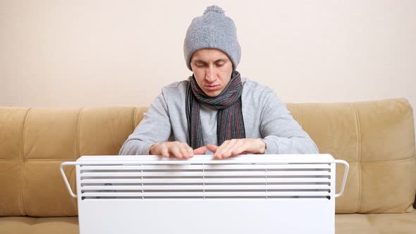 Sad Man with Scarf and Knitted Hat Warms Hands Above Heater