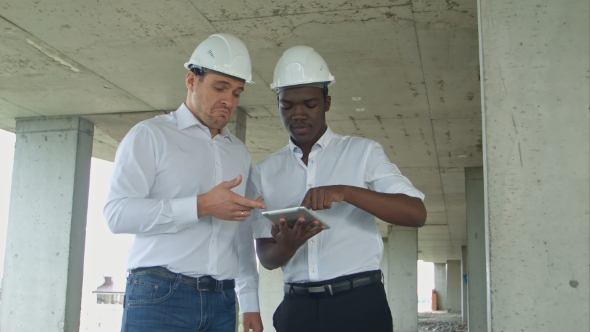 African American Engineer and Caucasine Architect Using Digital Tablet and Wearing Safety Helmets at alt
