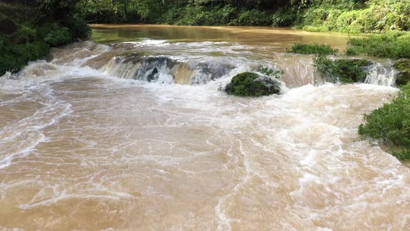 Still video of a small cascade showing the brown water running quickly with forest in the background