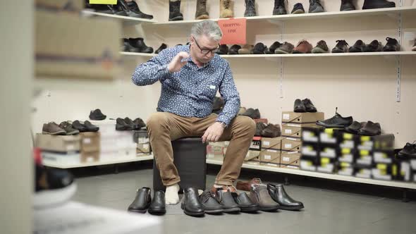 Wide Shot of Confused Senior Man Sitting in Shoe Store with Row of Boots Standing on the Floor