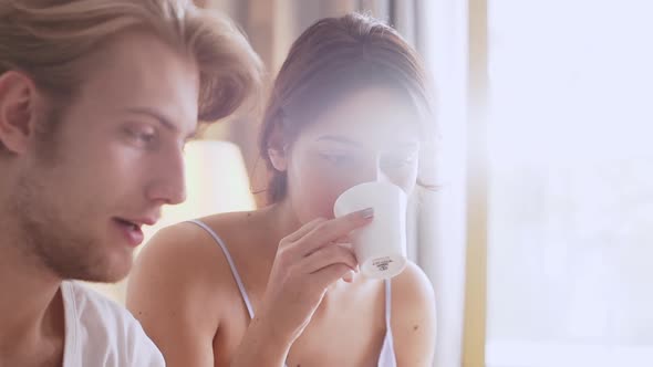 Attractive Caucasian Female Drinking Morning Coffee From Cup Sitting in Bed with Male Reading